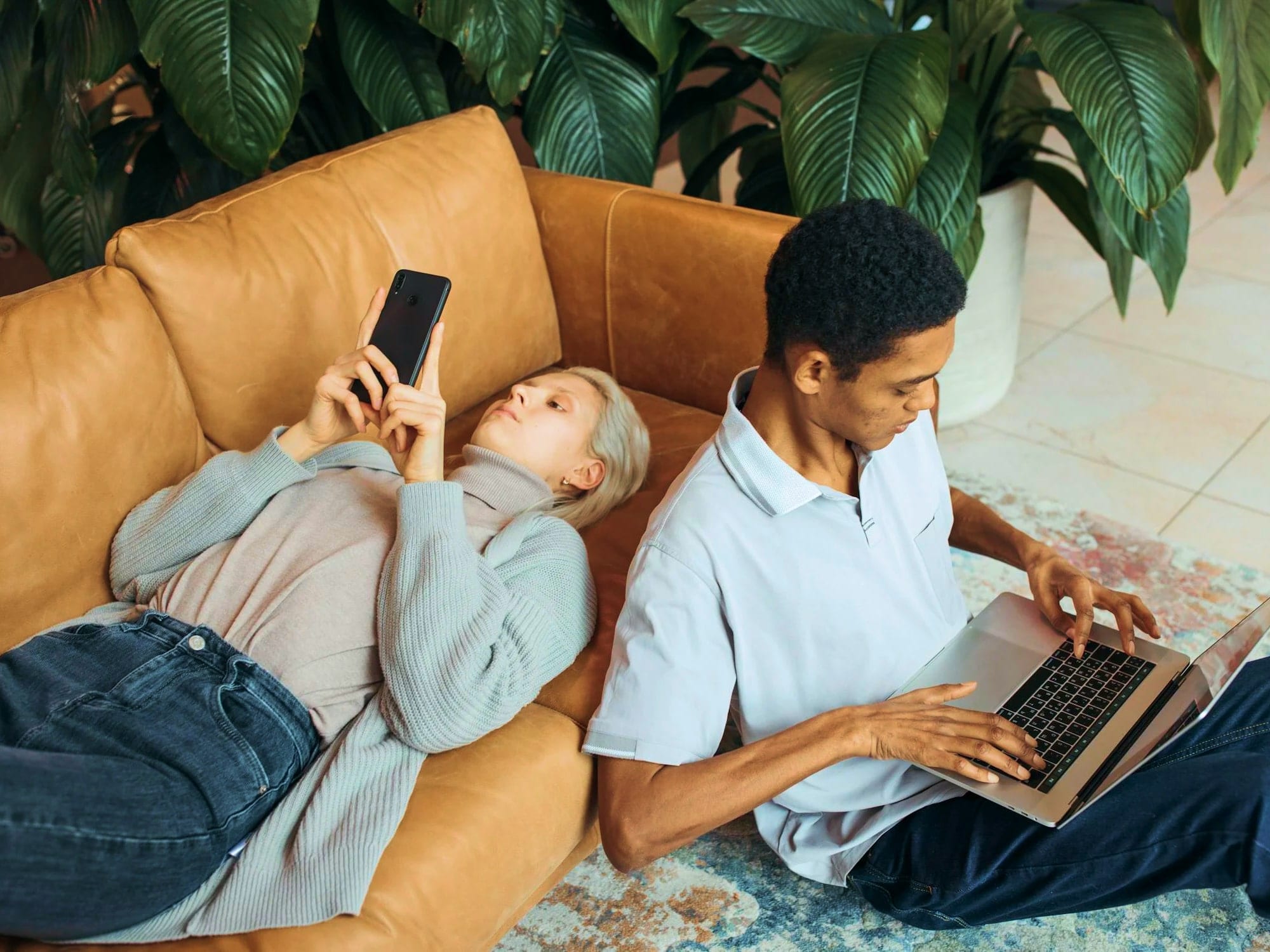 A woman lies on a couch using her phone while a man sits on the floor beside her working on a laptop. Large green plants are in the background.